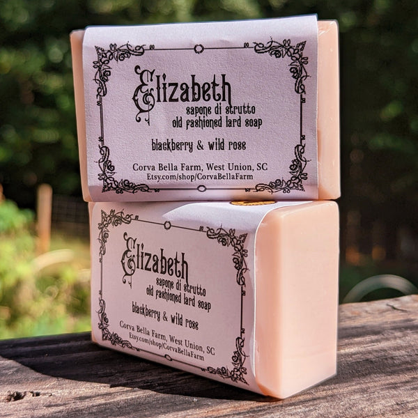 Two bars of lard soap with 'Elizabeth' written on the packaging, indicating a wild rose and blackberry scent, placed on a wooden surface with a natural background.